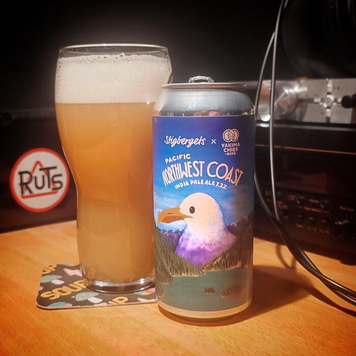 Ett glas öl bredvid en burk "Pacific Northwest Coast" India Pale Ale från Stigbergets och Yakima Chief Hops på ett bord.