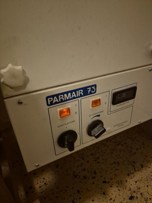 Ventilationssystem Parmair 73 med knappar för ventilation och eftervärme, monterat på vit panel mot fläckigt golv.