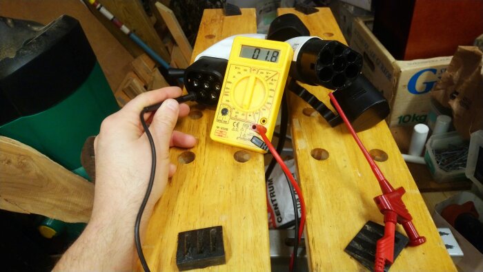 Digital multimeter mäter spänning vid laddkabel på arbetsbord. Hand håller sonder för att läsa värde på skärm. Verkstadsmiljö i bakgrunden.