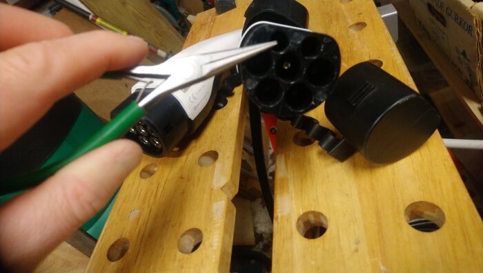 Tång och laddkontakt på träbord, tänger håller i grönt mätstift mot kontaktens stift, mäter resistans.