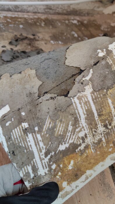 Plastmatta med grå massa och svart lim på undersidan, delvis frånskrapad betongyta, hålls i handen med handske.