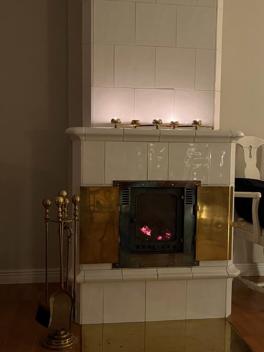 Vit kakelugn med brinnande glöd och guldiga spjäll, flankeras av verktyg i mässing mot en beige vägg.