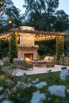 Uteplats med eldstad, skorsten och belysning under pergola, omgiven av gröna växter och blommor.