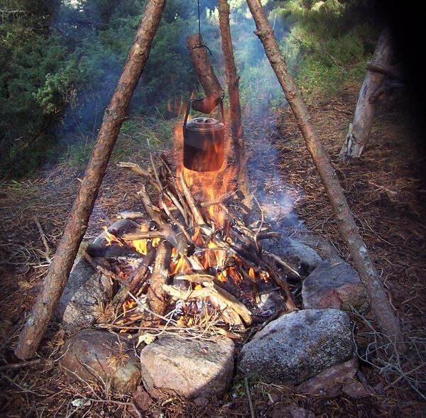 Lägereld omgiven av stenar med en hängande kaffepanna över elden, omgiven av natur och trädstammar.
