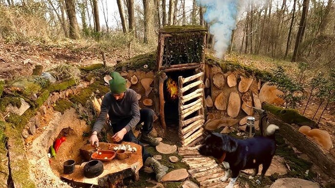 Man skapar en eldstad i naturen med jord runt omkring. En person förbereder mat medan en hund står intill. Rök stiger från en improviserad skorsten.
