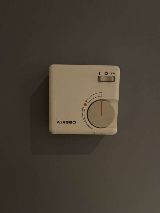Termostat från WIRSBO utan lock, med ratt för temperaturinställning, monterad på en mörk vägg.