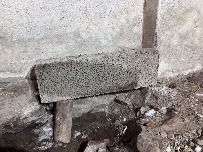 Betongplatta och timmer under en grundmur, omgiven av jord och små stenar.