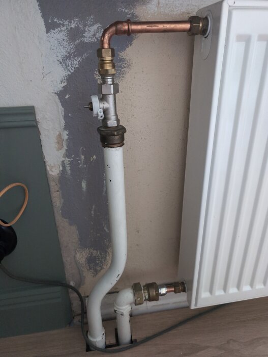 Radiator med kopparrör och termostatventil, visar anslutningen av inkommande och utgående vattenrör mot en vägg med flagnande färg.