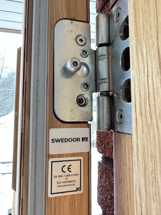 Dörrgångjärn med justeringsskruvar synliga, märkt med "SWEDOOR", monterat på en träram bredvid en murad vägg.
