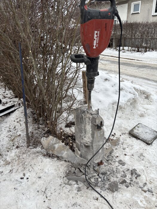Röd Hilti-borrmaskin bryter ner ett betongblock utomhus i snön, omgiven av buskar och en trottoar.