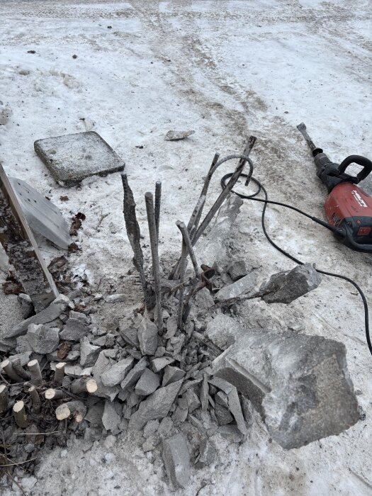 Raserad betongkonstruktion med utskjutande armeringsjärn på snötäckt mark, omgiven av stenar och en elektrisk bilningsmaskin.