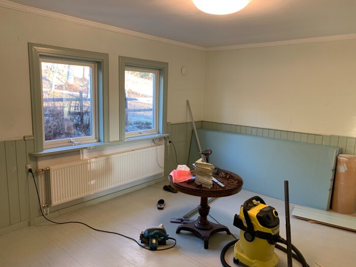 Rum under renovering med borttagen bröstpanel, två fönster, bord med verktyg, och en dammsugare.