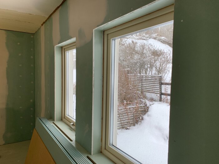 Rum under renovering med nylagd gips på väggarna och två fönster med utsikt över en snöig trädgård.