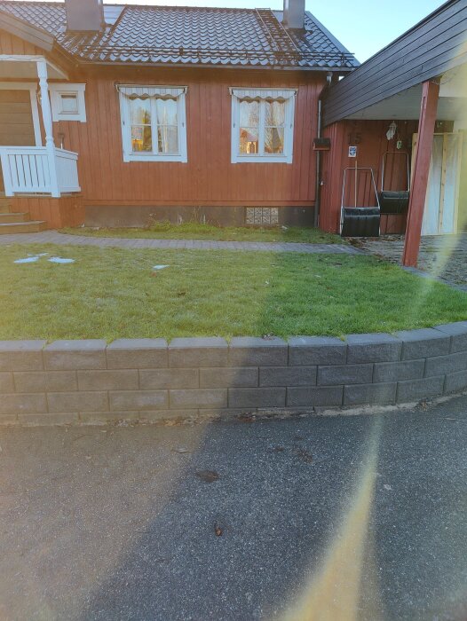 Rött hus med vita knutar, stenmur och gräsmatta framför, samt en svart ytterdörr under altan vid sidan. Solsken skapar linseringar över bilden.