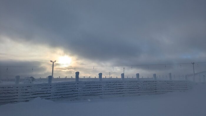 Snöigt vinterlandskap med molnig himmel, lågt stående sol, och ett snötäckt staket med jordstolpar i förgrunden.