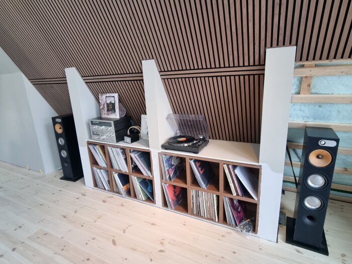 Hylla med vinylskivor, stereoanläggning och högtalare i ett halvfärdigt rum med träpanel på väggarna.