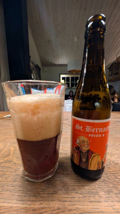 Ett glas med skummande mörk öl bredvid en flaska öl med etiketten "St. Bernardus Prior 8" på ett träbord i ett rum.