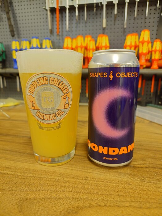 Ölglas med "Toppling Goliath Brewing Co." bredvid ölburk med texten "Shapes & Objects" i en verkstadsmiljö.