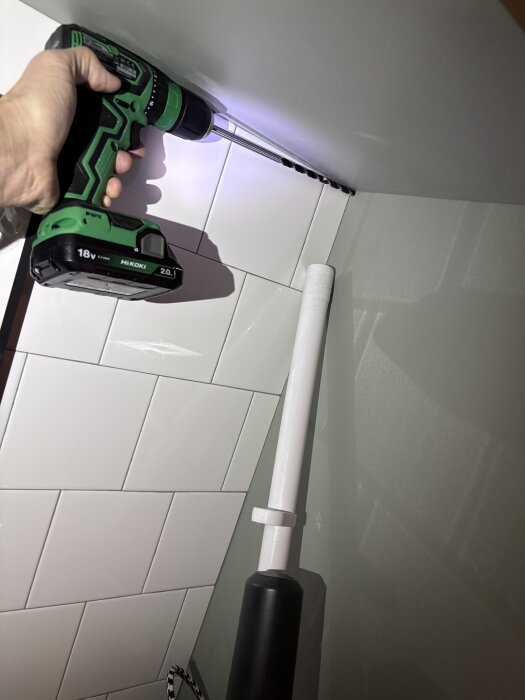 Kakelvägg med grönt elverktyg borrande i hörn, vitt rör bredvid.