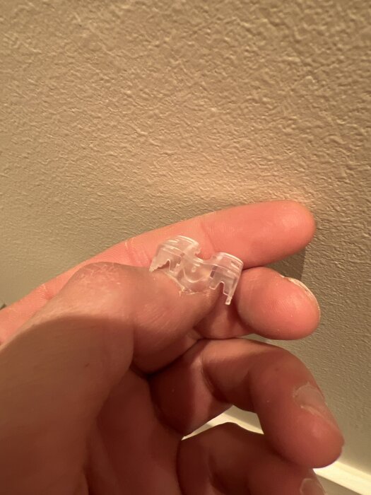 En hand håller en transparent plastbit som används för nedtryckning av kablar mot en beige vägg.