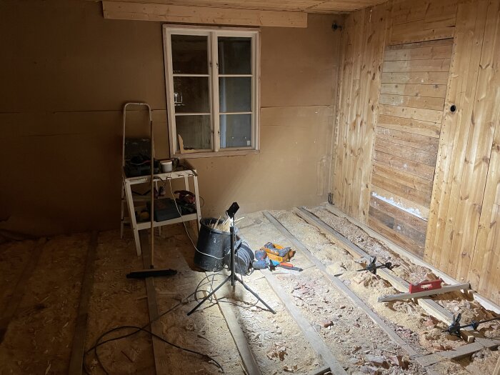 Renovering av kök pågår med borttaget golv och verktyg som stege, såg och skruvdragare synliga. Fönster och träpanel finns i rummet.