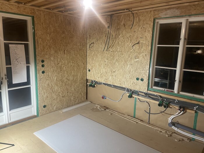 Renovering av kök med uppsatta OSB-skivor och exponerade rör för el och VVS, samt ett fönster och en dörr med anslag på väggen.