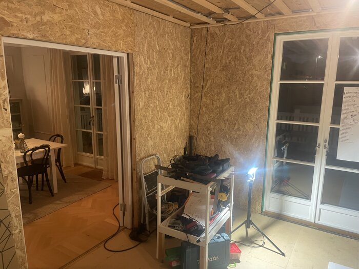 Rum under renovering med verktygshylla, arbetslampa och osb-väggar, angränsande till matsal med bord och stolar.