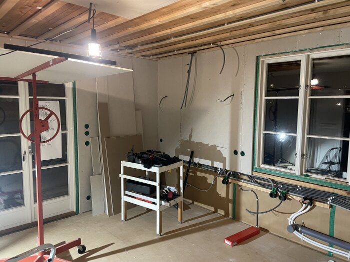 Renovering av kök med verktyg och byggmaterial, hängande taklampa, synliga elkablar på en vägg och fönster till höger.