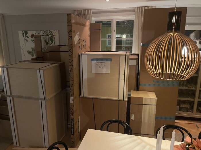 Kartonger staplade i ett vardagsrum, redo för köksrenovering, med en modern lampskärm och ett matbord i förgrunden.