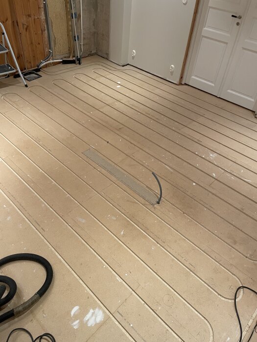 Bild av ett rum under renovering med golvvärmeplattor installerade. Vita väggar och en dörr syns i bakgrunden. Byggmaterial ligger på golvet.
