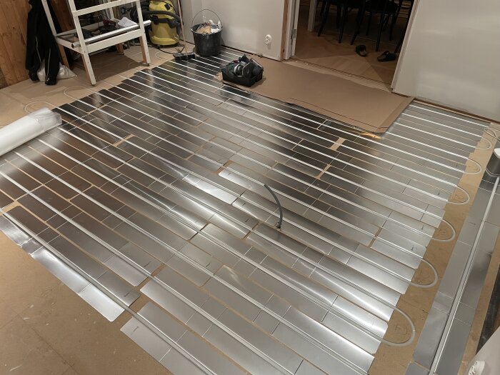 Golvläggning med synliga värmefördelarplåtar och rör för golvvärme i ett rum under renovering, med verktyg och utrustning i bakgrunden.