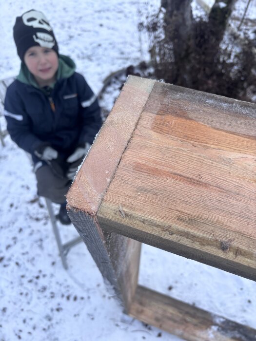 Träram byggd för varmbänk syns i närbild, en pojke med mössa sitter i snön intill.