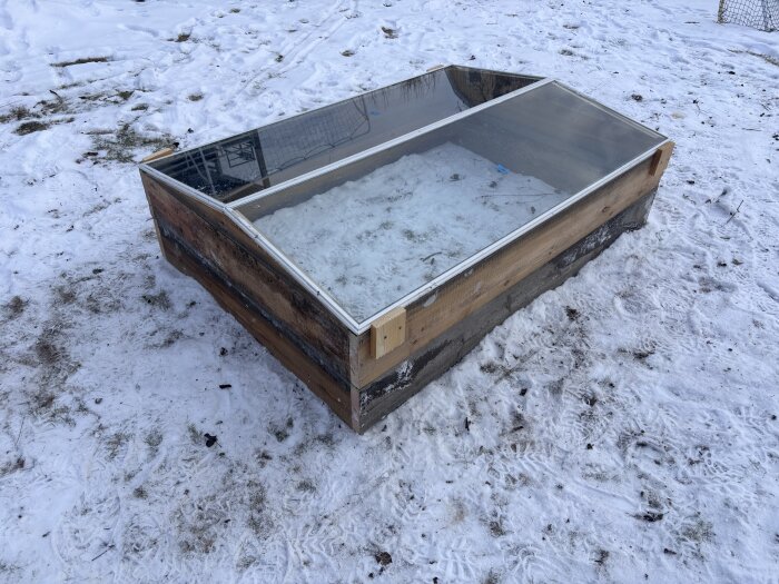 Träram med glaslock för varmbänk, placerad på snötäckt mark. Används för att odla tidiga grödor som spenat och sallad.