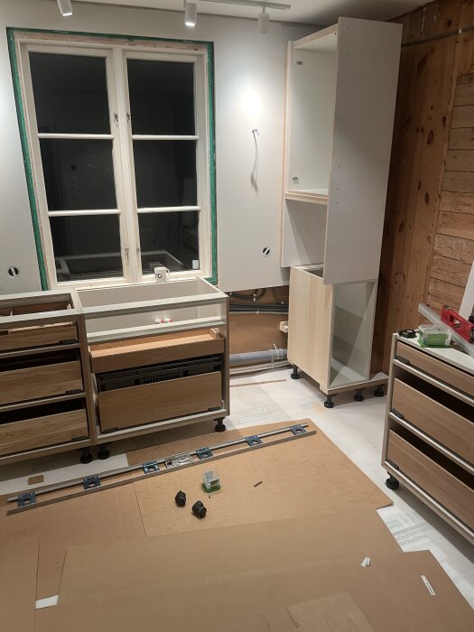 Kök under konstruktion med halvfärdig stomme och överskåp, verktyg och material på golvet framför ett obehandlat fönster.