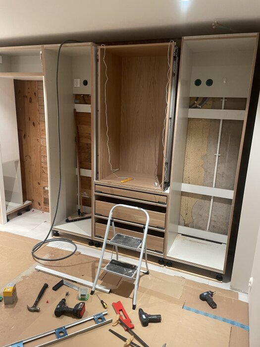 Påbörjad köksrenovering med öppna skåp och verktyg utspridda på golvet, stege och material syns i förgrunden.