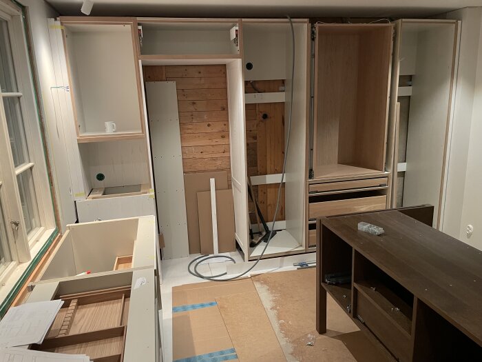 Nytt kök under konstruktion med halvfärdiga skåp, bänkar och träpanel på väggen, samt verktyg och material spridda i rummet.