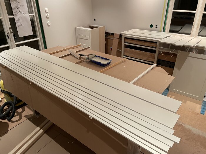 Byggmaterial och verktyg påbörjar köksmontering, vita hyllplan och lådor är delvis installerade i ett oavslutat köksutrymme.