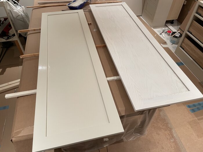 Två vita köksluckor ligger på ett bord, omgivna av byggmaterial, i ett köksrenoveringsprojekt.