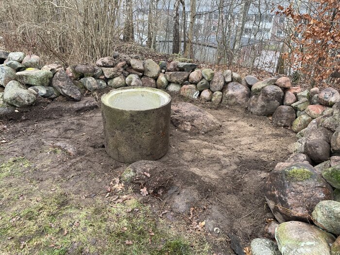 Stenmur i U-form med en stencirkel i mitten på en grusad yta, omgiven av träd och buskar i bakgrunden.