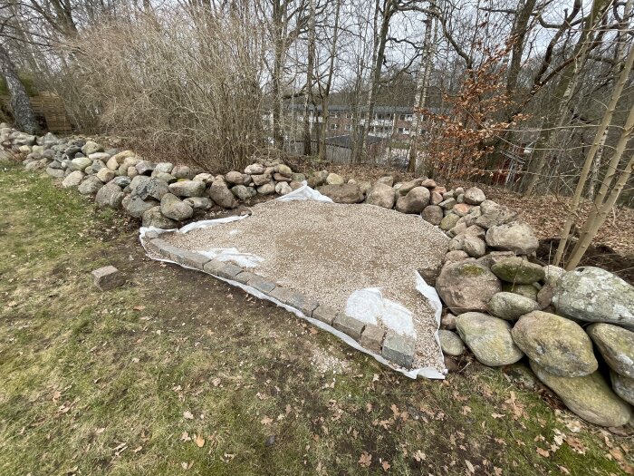 Stenmursomgärdad grillplats med singel på marken, omgiven av träd och buskar i trädgård.