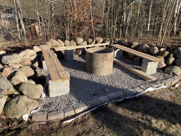 Grillplats med grusbelagd yta omgiven av stenmur. Två träsitsar och eldstad i mitten, beläget i skogsmiljö.