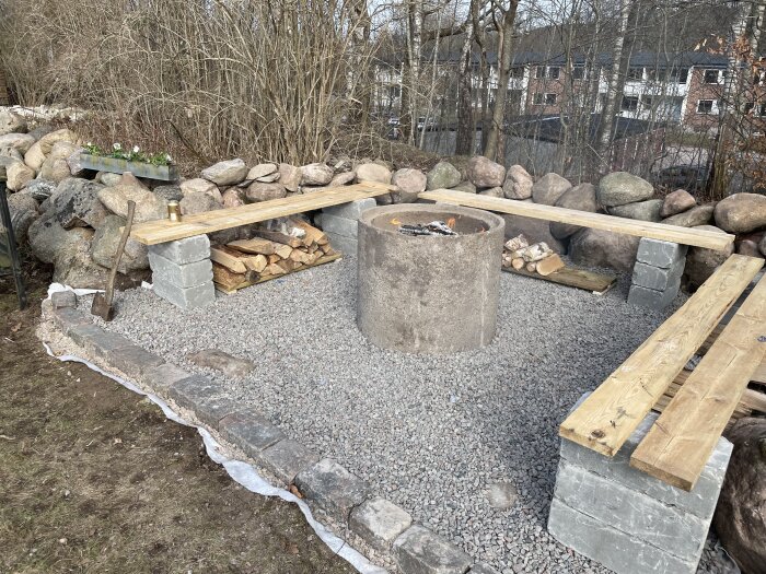 Grillplats med rund eldstad av betong, omgiven av träbänkar och stenmurar, samt ved och stenar på grusunderlag i en trädgårdsmiljö.