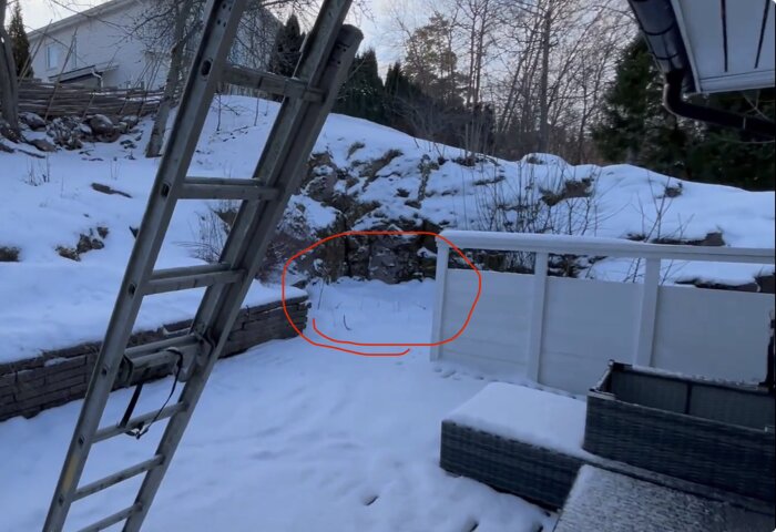 Snötäckt bakgård med stenmurar och en stege, omgiven av träd och vinterlandskap. En markering visar platsen för en grillplats.