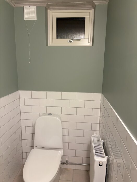 Vit toalettsits, gröna väggar, vitt kakel och radiator i nytt badrum.
