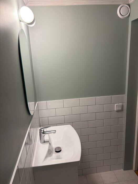 Badrum med gröna väggar, vit kaklad vägg, handfat med kran och rund spegel under en vägglampa.
