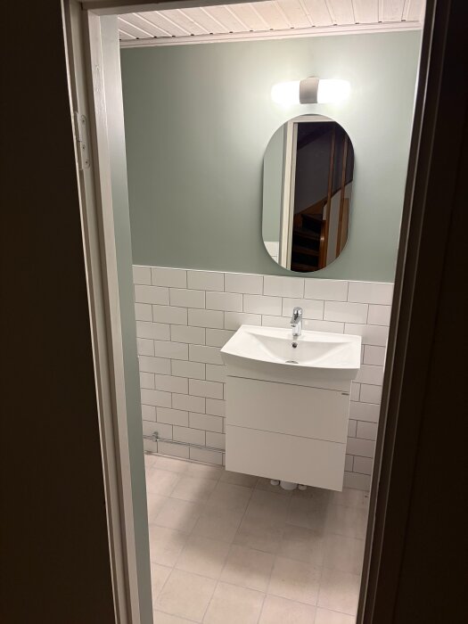 Badrum med ljusgrön vägg, vit kakelpanel, ovalt spegel och vit handfat med kommod.