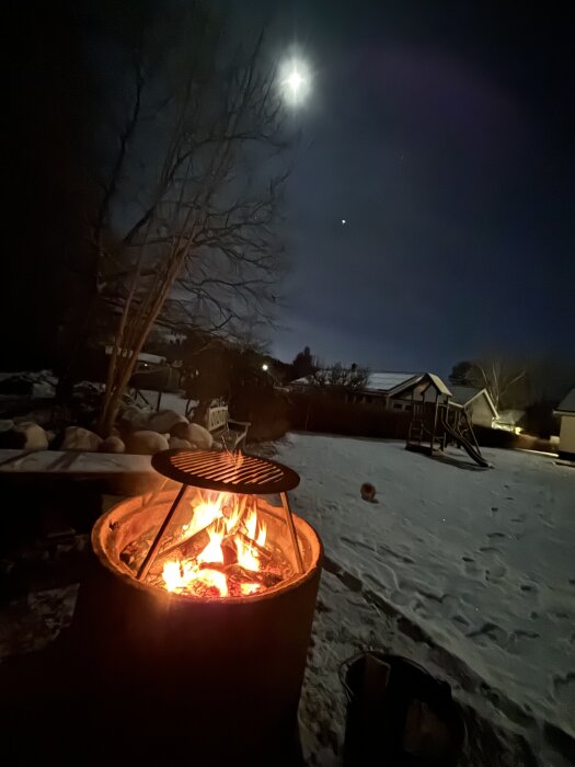 Upplyst grillplats med glödande eld, snötäckt bakgård och måne i natthimlen.