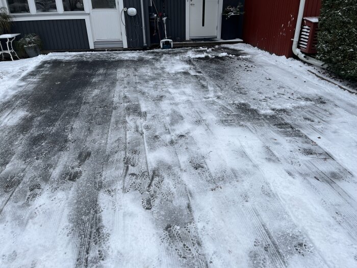 Snötäckt betonguppfart framför ett hus med röd fasad och fönster, snöskovel synlig vid dörren och fotspår i snön.