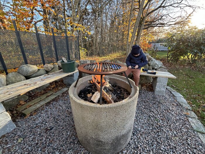 En utomhusgrillplats med en stenmur i U-form, en eldstad med grillgaller och en person som sitter på en bänk bland höstlöv.