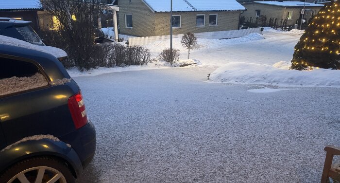 Snöbeklädd uppfart och bil, kvällstid, med upplyst gran i bakgrunden. Snön är fluffig och lätt, typisk för vintersäsongen.